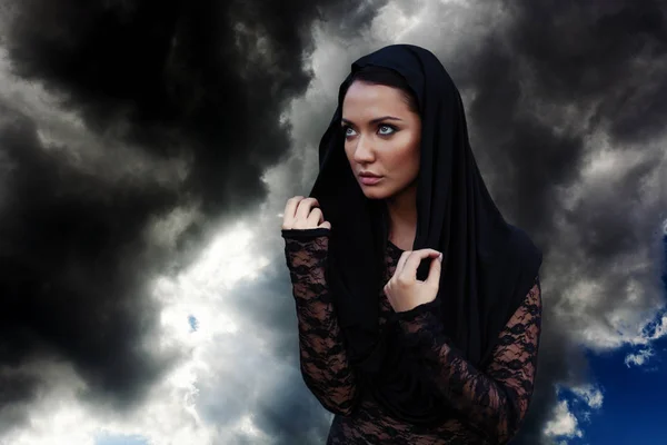 Mulher bonita nova com o cabelo preto e no capuz preto e blusa transparente preta no fundo do céu — Fotografia de Stock