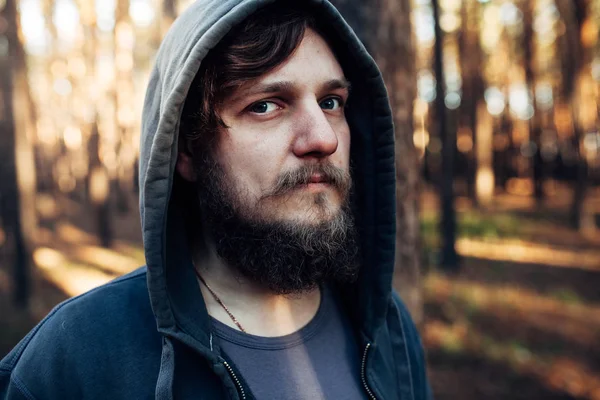 close up portrait of a bearded hipster tourist in gray hood man in the sunlight woods forest