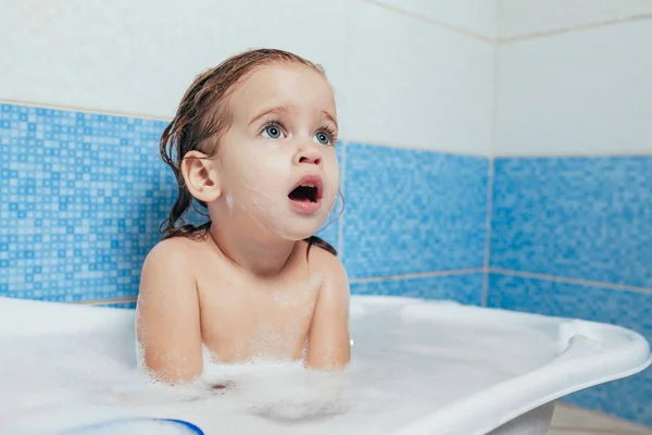 Fun cheerful happy toddler baby taking a bath playing with foam bubbles. Little child in a bathtub. Smiling kid in bathroom on blue background. Hygiene and health care. Stock Photo