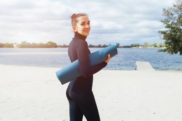 An athletic woman is standing with her back to the camera on the lake shore and is holding a yoga mat.