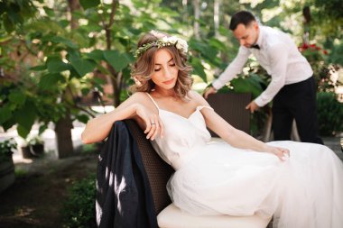 A beautiful bride in a white wedding dress and wreath sits on a chair next to the groom, resting and preparing for a happy family life clipart