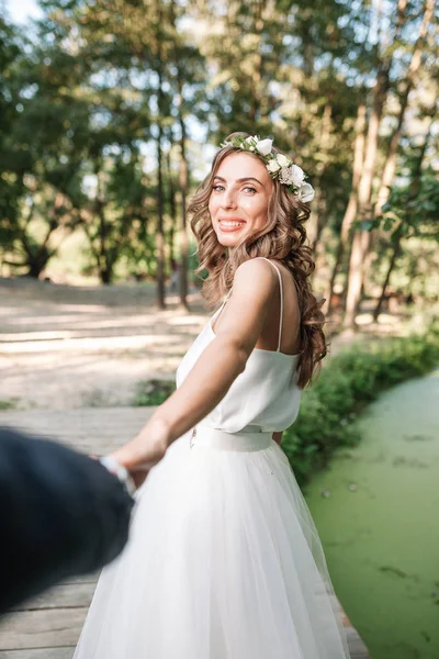 Следуй за моей концепцией любви. Привлекательная молодая женщина, одетая в белое свадебное платье, держит за руку своего парня и идет по прекрасному саду к счастливому будущему — стоковое фото