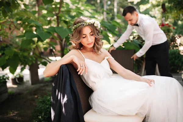 Eine schöne Braut in weißem Brautkleid und Kranz sitzt auf einem Stuhl neben dem Bräutigam, ruht sich aus und bereitet sich auf ein glückliches Familienleben vor — Stockfoto