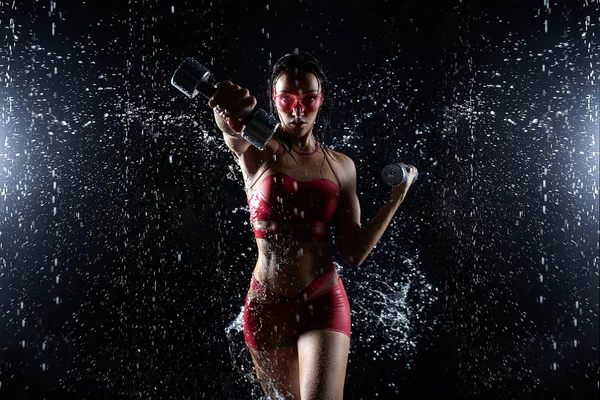 Krásná mladá dívka v červených sportovní představuje s činkami v aqua studio. Kapky vody šíří o její vhodnosti tělo. Dokonalá postava na pozadí postříkání vodou — Stock fotografie