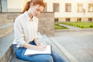 Genç ve güzel bir öğrenci not defterine bir kompozisyon yazıyor. Merdivenlerde oturuyor. Kızıl saçlı, çilli kız.