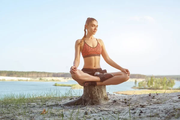 Young beautiful girl dressed in sportswear and sneakers sits in the lotus position in the summer in the park against the lake — Stock Photo, Image