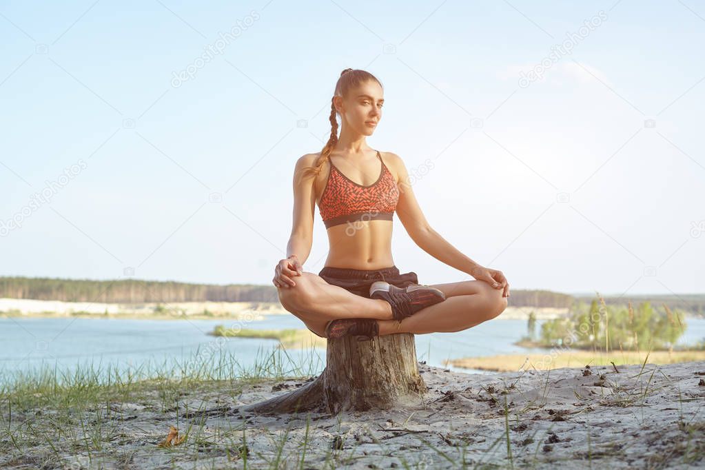 Young beautiful girl dressed in sportswear and sneakers sits in the lotus position in the summer in the park against the lake.