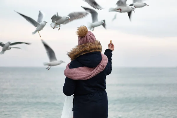 冬服で一人の女の子はドックの上に立って 彼らの手からカモメを供給します 冬の海と鳥 寒さで海で認識できない人 — ストック写真