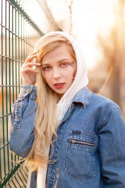 Yüzünde geçici olarak boyanmış çiçekler olan genç bir kadın. Uzun sarı saçlı genç kız kot ceket ve kapüşon giyiyor.