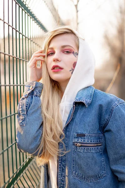 Mulher jovem com flores pintadas temporárias no rosto. adolescente com cabelo loiro longo usar jaqueta de jeans e com capuz — Fotografia de Stock