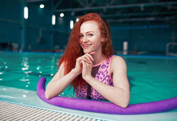 Hermosa chica pelirroja joven en un traje de baño de moda en el estilo de los años 80 se encuentra con los fideos en sus manos en la piscina cubierta — Foto de Stock