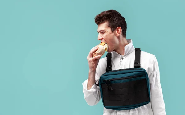 Genç şık erkek işçi göğüs teçhizat çanta ile beyaz bir arka plan üzerinde lezzetli sulu hamburger yiyor — Stok fotoğraf