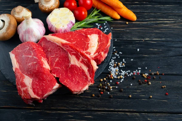Drei Stücke saftiges rohes Rindfleisch mit Gemüse auf einem Steinschneidebrett auf schwarzem Holztischhintergrund. — Stockfoto