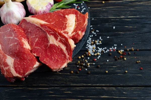Drei Stücke saftiges rohes Rindfleisch mit Gemüse auf einem Steinschneidebrett auf schwarzem Holztischhintergrund. — Stockfoto