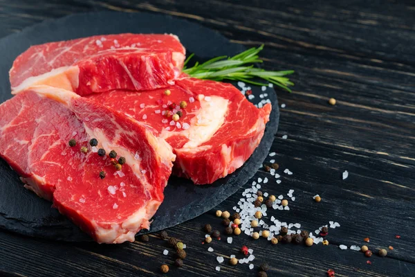 Tres piezas de jugosa carne cruda en una tabla de cortar de piedra sobre un fondo de mesa de madera negro . —  Fotos de Stock