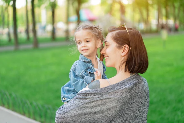 A little Caucasian girl dressed in a denim jacket sits in her mother's arms. Mom and daughter gulf in the park on a hot summer day — Stock Photo, Image