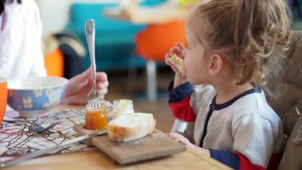 Kleines kaukasisches Mädchen, Kind, isst leckeres Brot und Marmelade an einem Tisch in einem Café mit ihrer Mutter. — Stockvideo