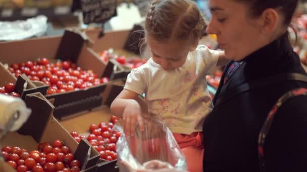 Mamá con su hijita en brazos recogiendo tomates en un supermercado. Compradores en la tienda . — Vídeo de stock