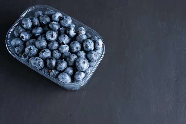 Tasty juicy raw blueberries in a plastic container on a black dark background. — Stock Photo, Image