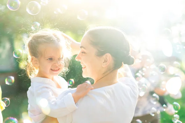 Little  lovely girl and her mother having fun with soap bubbles on blurred nature background. Outdoors at the daytime with bright sunlight. — Stock Photo, Image