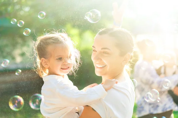 Little  lovely girl and her mother having fun with soap bubbles on blurred nature background. Outdoors at the daytime with bright sunlight. — Stock Photo, Image