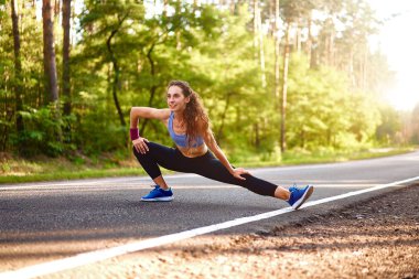 Genç kadın koşudan önce orman yolunda ısınır. Egzersizler çalıştırmak için uzanan çekici kız. Güzel kıvırcık sportif kız fitness açık yapıyor 