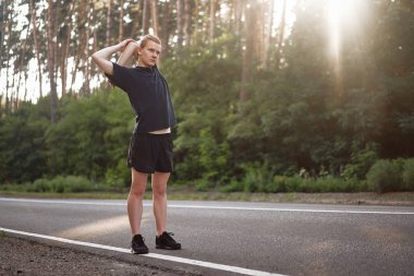 Genç kıvırcık adam koşudan önce orman yolunda ısınır. Egzersizler için esnenerkekler. Yakışıklı 20s athlet fitness açık yapıyor 