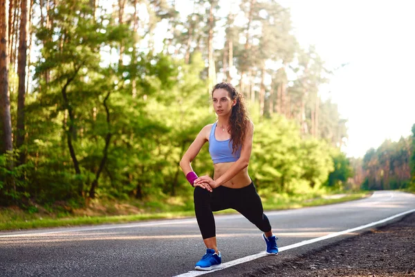 Young woman warming up at forest road before jogging. Attractive girl stretching for running exercises. Beautiful curly sporty girl doing fitness outdoor — Stock Photo, Image
