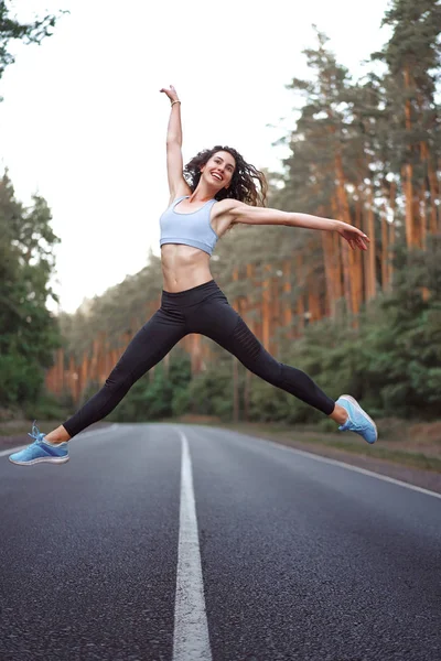 Young beautiful brunette girl jumping in twine with background  asphalt road and pine forest — Stock Photo, Image