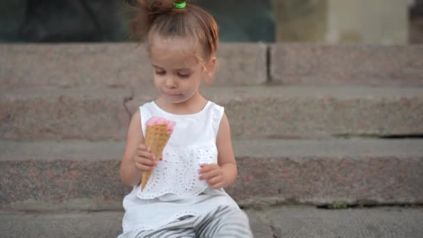 Cute caucasian little girl enjoys ice cream in a waffle cone walking in the park. The child eats bright ice cream — Stock Video