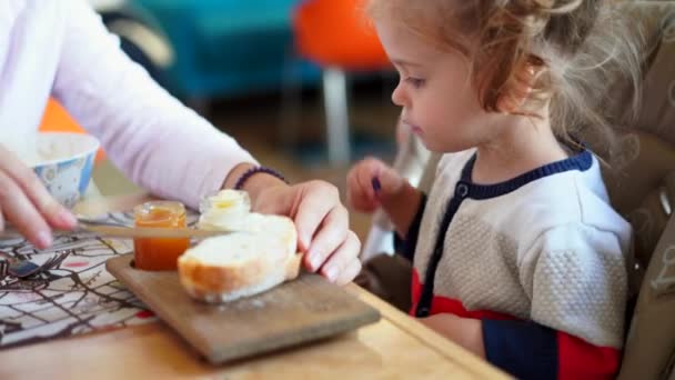 Menina caucasiana, criança, comendo delicioso pão e geléia em uma mesa em um café com sua mãe . — Vídeo de Stock