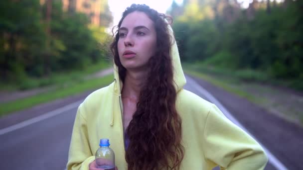 Bela jovem caucasiana de pé na estrada da floresta de asfalto Bebe água de garrafa de plástico depois de correr — Vídeo de Stock