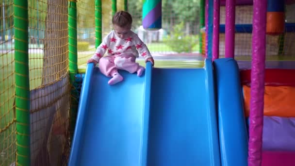 Маленькая девочка спускается от пластиковых слайдов к мячам на детской площадке — стоковое видео