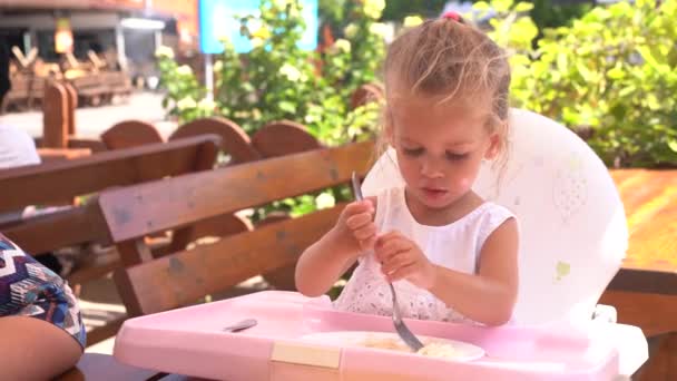 Linda niña caucásica comiendo espaguetis en la mesa sentado en el asiento del niño restaurante al aire libre . — Vídeos de Stock
