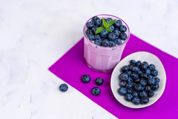 Tasty fresh blueberry yoghurt shake dessert in glass standing on white table purple napkin background. — Stock Photo, Image