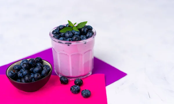 Tasty fresh blueberry yoghurt shake dessert in glass standing on white table purple napkin background. — Stock Photo, Image