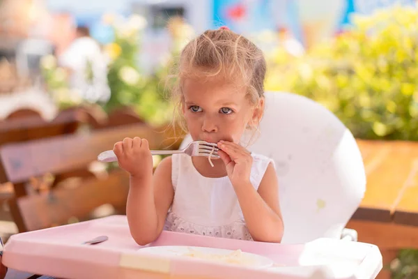 Schattige kleine Kaukasische meisje eten spaghetti op tafel zitten in kinderzitje outdoor restaurant. — Stockfoto