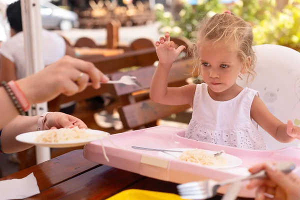 Schattige kleine Kaukasische meisje eten spaghetti op tafel zitten in kinderzitje outdoor restaurant. — Stockfoto