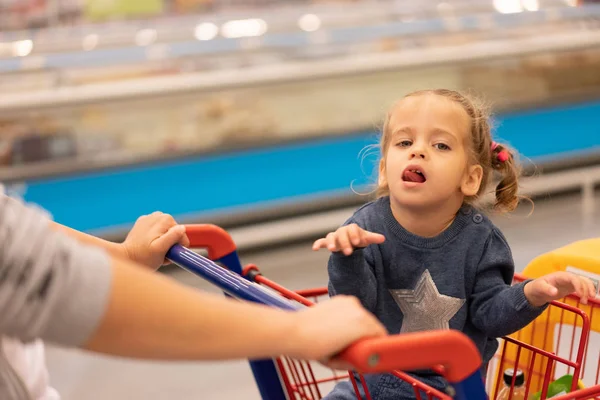 Klein meisje zit Shopping Trolley in de buurt van winkels. — Stockfoto