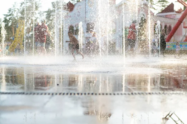 Niños felices irreconocibles se divierten jugando en la fuente de agua seca de la ciudad en el caluroso día de verano —  Fotos de Stock