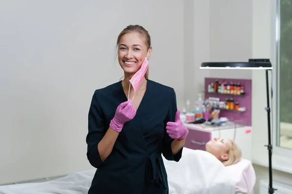 Cosmetology cabinet client lies on couch. Beautician stands in pink medical mask and smiles. — Stock Photo, Image