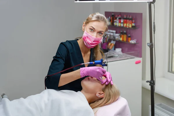 Cosmetology cabinet client lies on couch. Beautician applies marking on eyebrows. — Stock Photo, Image