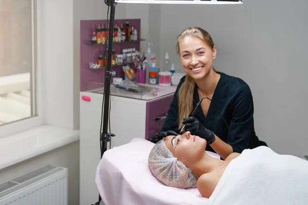 Cosmetology cabinet client lies on couch. Beautician applies marking on eyebrows. — Stock Photo, Image