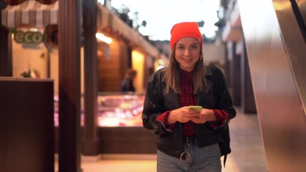 Una joven hermosa chica caucásica camina por el centro comercial con chaqueta de cuero negro y jeans se ve en la pantalla de su teléfono . — Vídeos de Stock