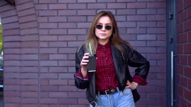 Jong mooi meisje student staat in de buurt baksteen muur in de herfst in zonnebril houdt koffie in haar handen en lacht. — Stockvideo