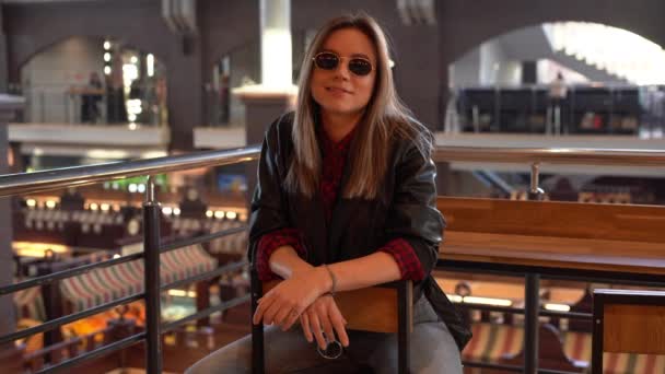 Uma bela jovem europeia moderna com cabelos brancos em uma jaqueta de couro preto e jeans senta-se café interior em um belo dia de outono . — Vídeo de Stock