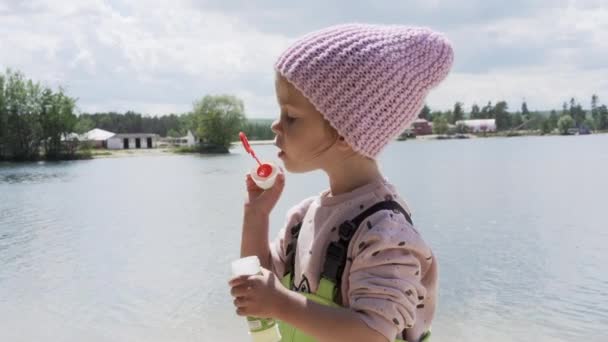 Küçük kız dışarıda sabun köpüğü üflüyor. Sonbahar pembe şapkası göl kenarında. — Stok video