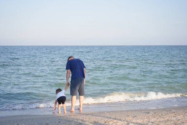 Baba oğlunun elini tutuyor. Baba Çocuk deniz tatilinde birlikte vakit geçiriyor. Küçük çocuk kumsalda yürüyor. Babalar günü. Tek çocuklu bir aile. Babanızla mutlu bir çocukluk. Tam uzunluk