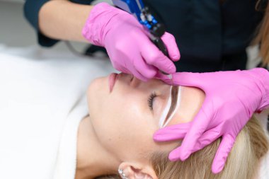 Kozmetik dolabı müşterisi kanepede yatıyor. Güzellik uzmanı kaşlara kalıcı mürekkep uygular. Kalıcı kaş makyajı prosedürü. Kapatın. Güzellik endüstrisi Pembe eldivenler