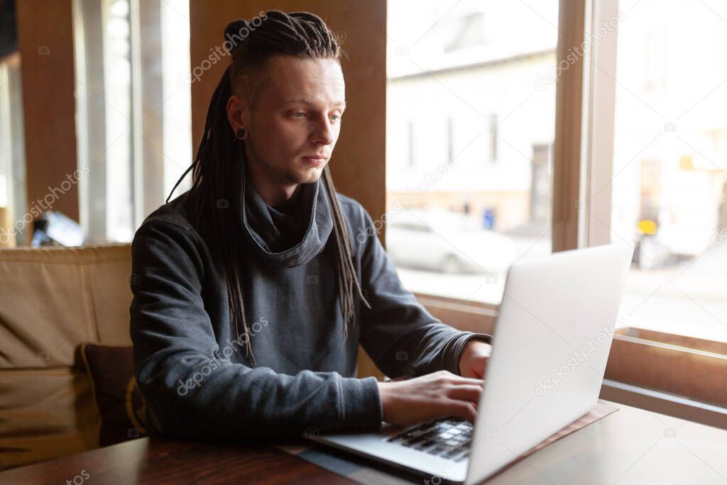 Young Businessman with dreadlock having doing his work in cafe near the big window with laptop. Youth culture. Neformal guy. Remote works. IT specialist